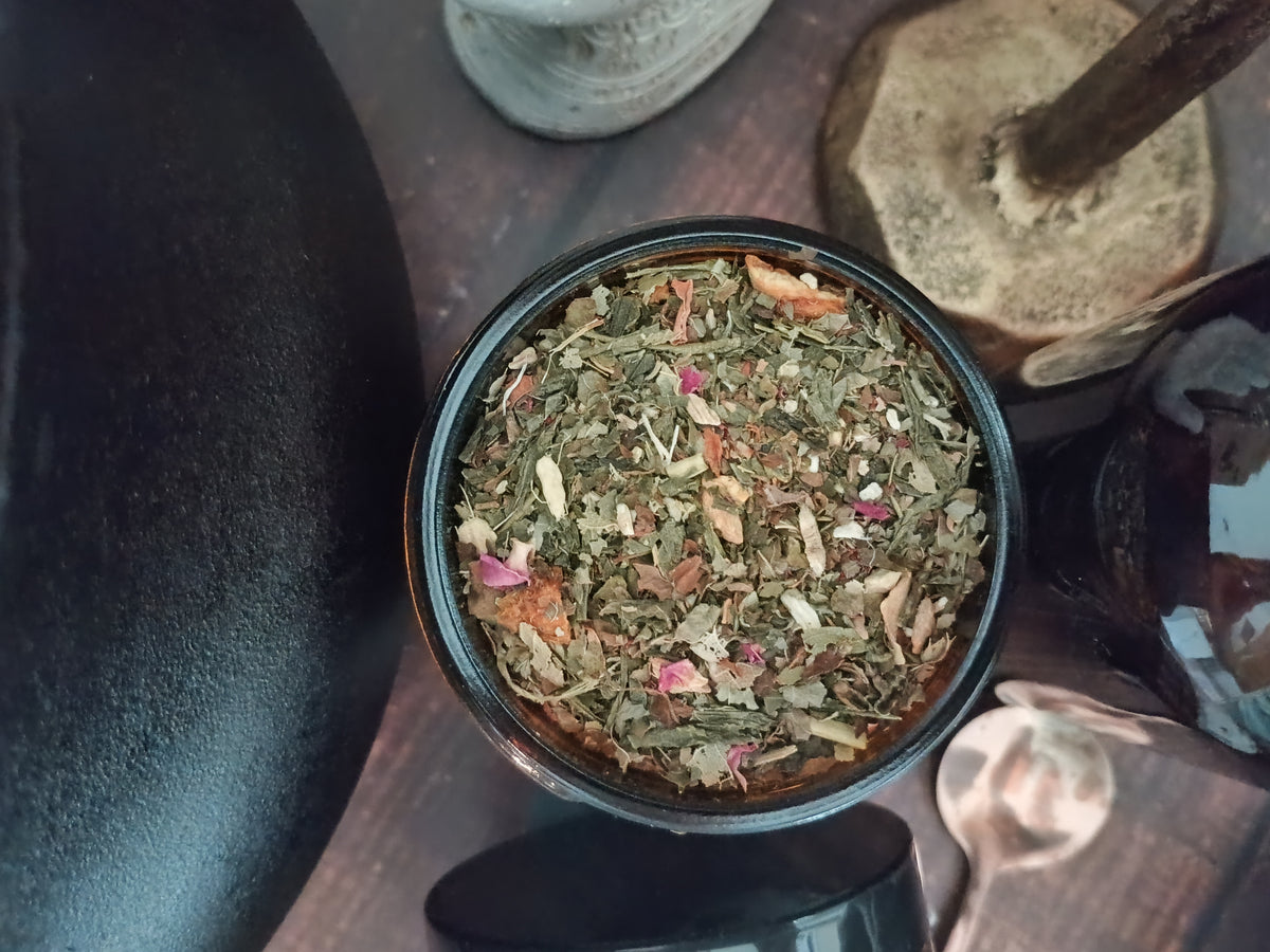 An overhead close-up of loose-leaf herbal tea in a small dark round container, with dried green leaves, orange pieces, and pink flower petals visible on a dark wooden surface.