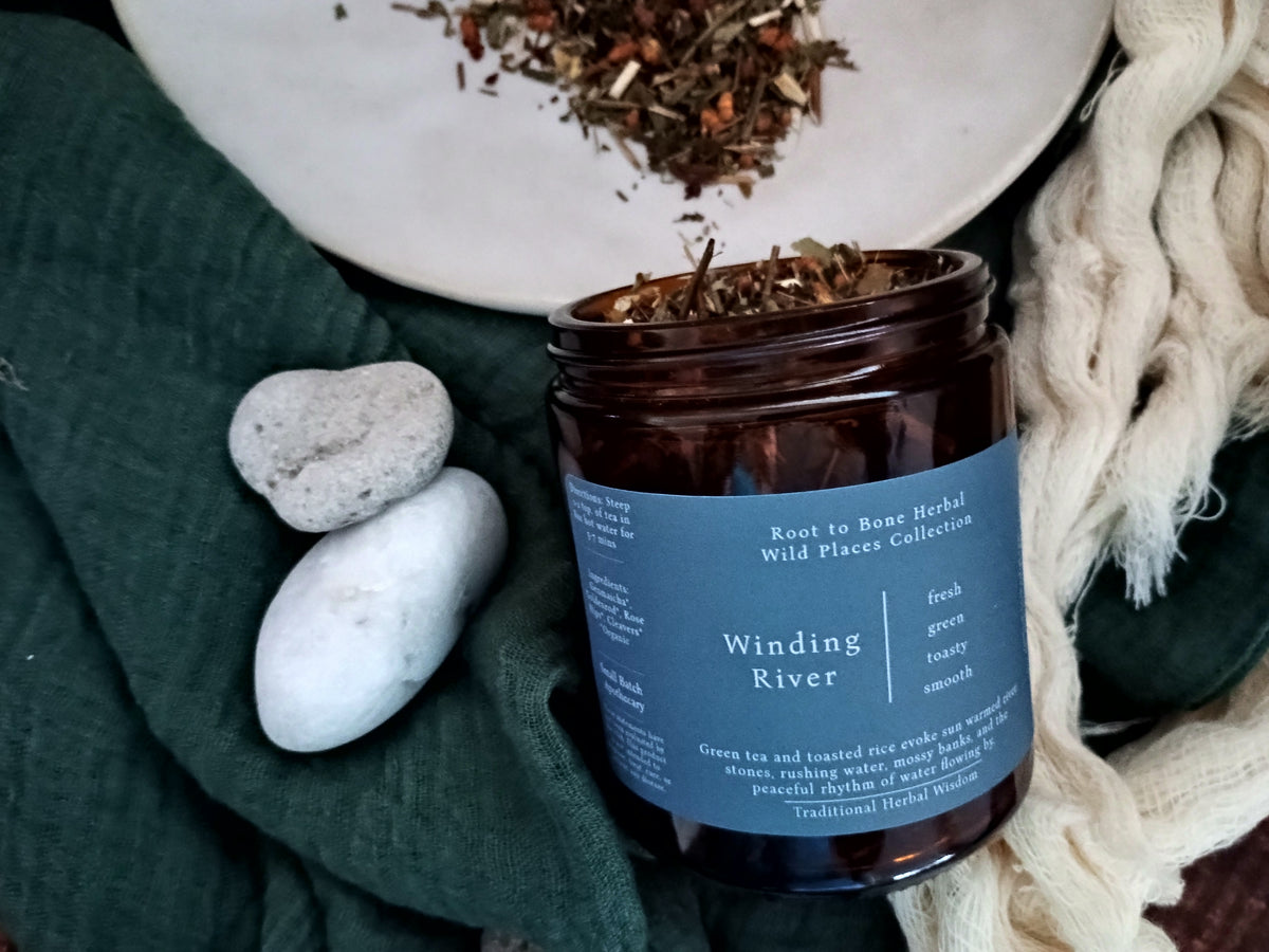 An amber glass jar of Winding River Herbal Tea with a slate blue label, styled on a dark green fabric with white stones and a cream knit blanket.
