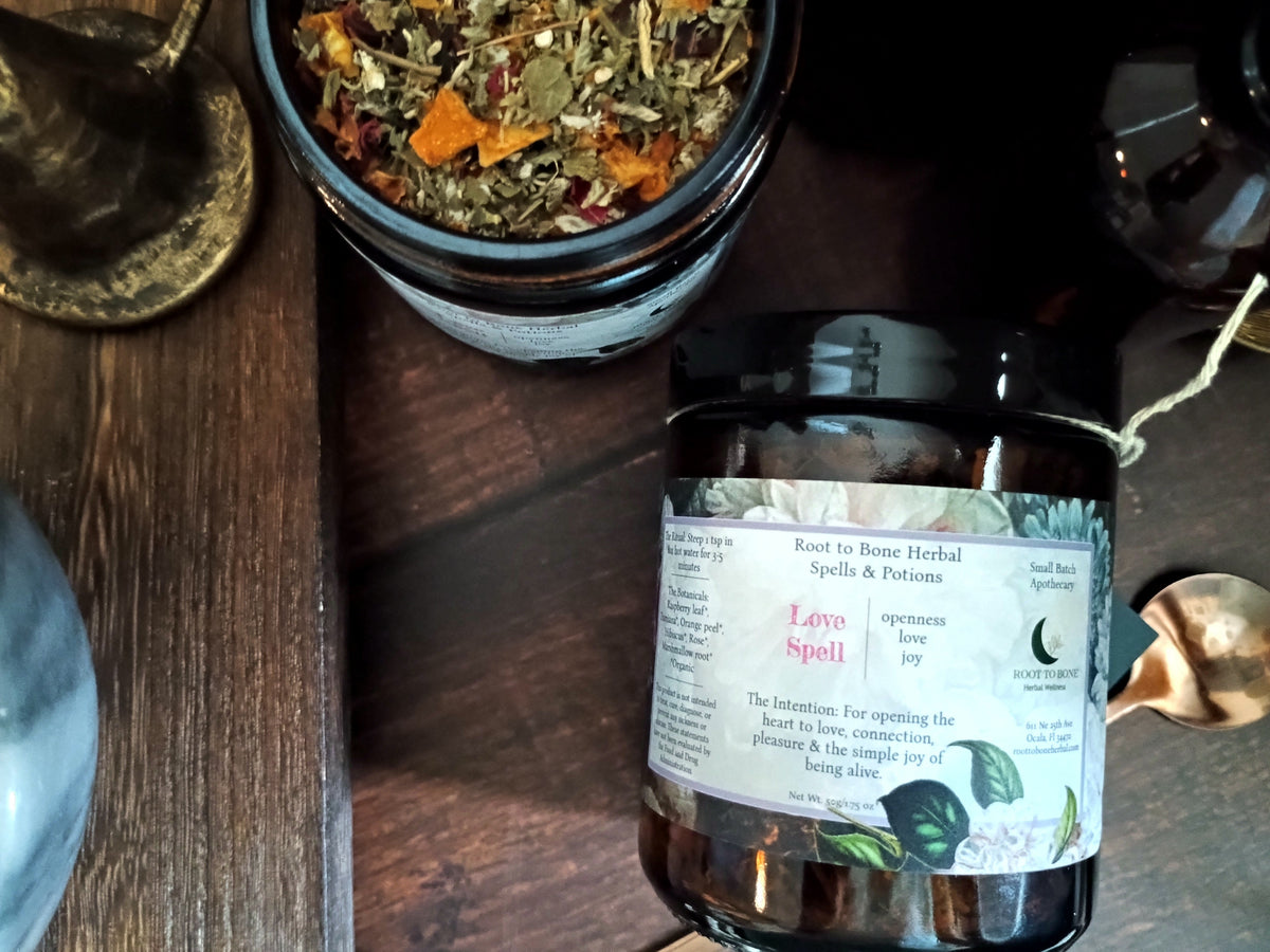 A dark amber glass jar labeled 'Love Spell' with a white label, placed on a dark wooden surface next to an open jar of loose herbal blend. The scene includes rustic props like a brass spoon and a dark bottle in a styled tabletop setting.