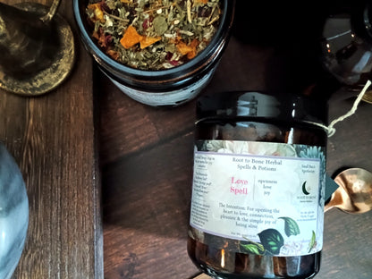 A dark amber glass jar labeled 'Love Spell' with a white label, placed on a dark wooden surface next to an open jar of loose herbal blend. The scene includes rustic props like a brass spoon and a dark bottle in a styled tabletop setting.