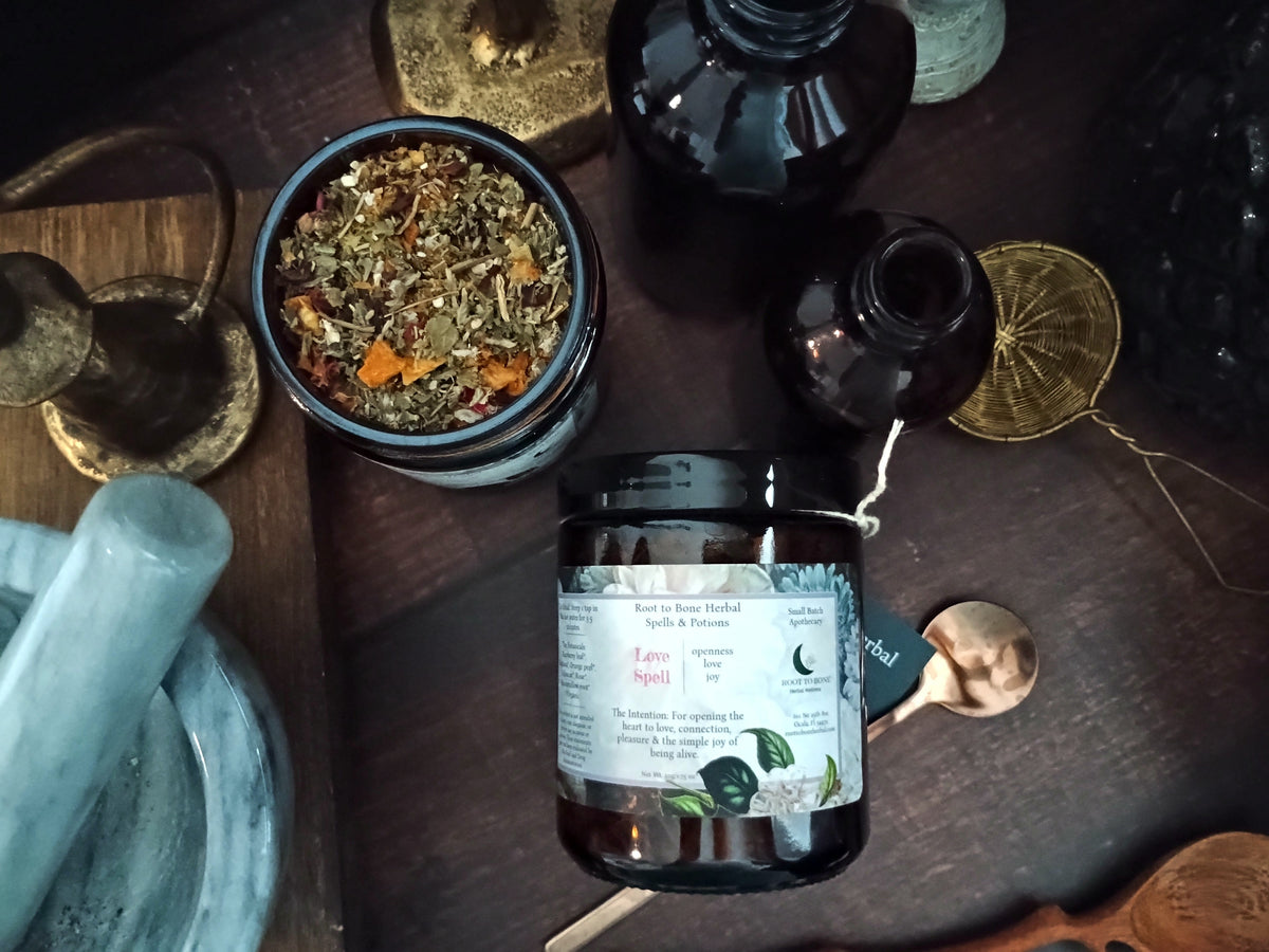 A jar of Love Spell herbal loose leaf tea is displayed on a dark wooden table alongside a bowl of loose herbal tea, dark glass bottles, a brass spoon, and a wooden spoon.