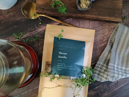 Tea package labeled 'Throat Soothe' on a wooden surface with herbs and a teapot.