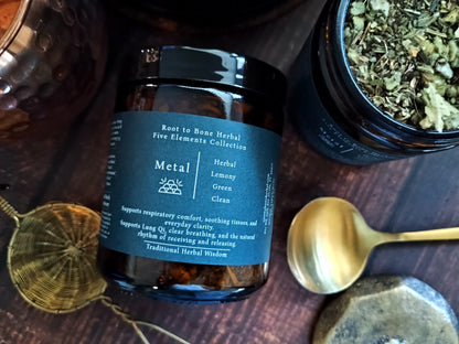 A dark amber glass jar of Root to Bone Herbal Five Elements Collection 'Metal' tea, surrounded by herbal ingredients in a jar, a brass spoon, and a brass strainer on a dark wooden background.