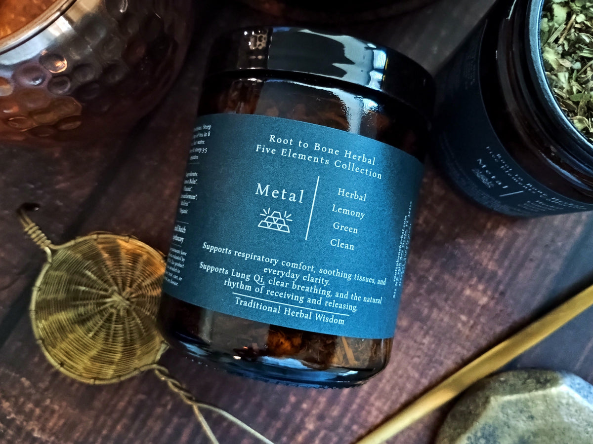 A dark amber glass jar of 'Metal' herbal tea from the 'Root to Bone Herbal Five Elements Collection' with a dark blue label, styled on a dark wooden surface with a brass strainer and other jars.