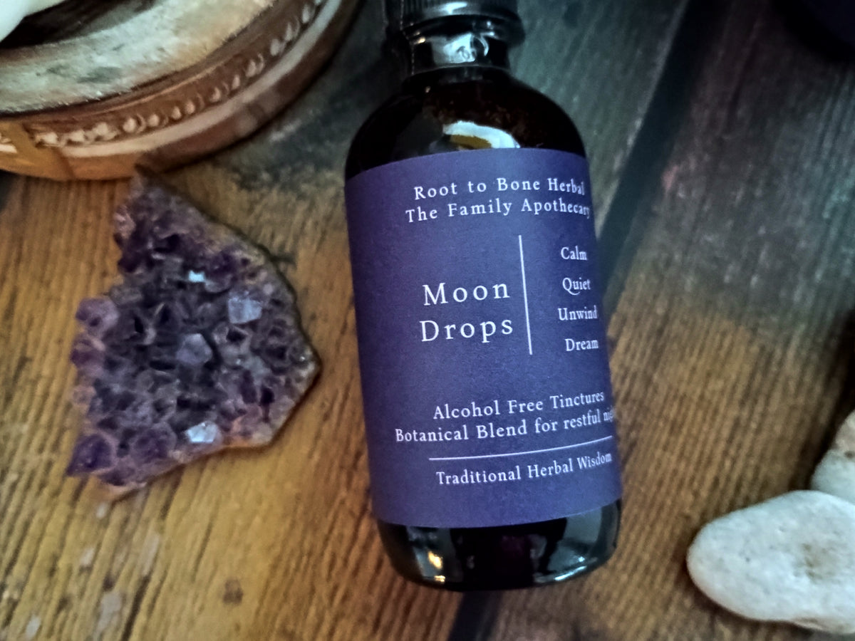 A dark glass bottle of Root to Bone Herbal Moon Drops with a purple label, styled on a wooden surface with an amethyst cluster and crystals.