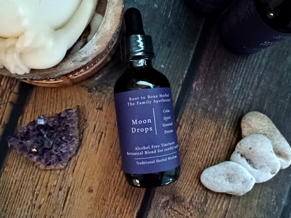 A dark amber glass dropper bottle labeled 'Moon Drops' by Root to Bone Herbal, placed on a rustic wooden surface with decorative stones and a bowl of white wax around it.