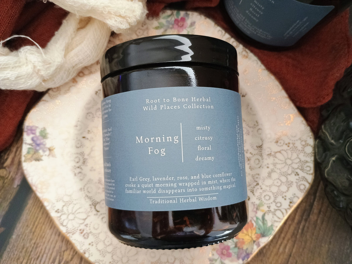 A dark amber glass jar of Root to Bone Herbal 'Morning Fog' loose leaf tea featuring a light blue label and black lid, styled on a floral ceramic plate with rustic wool and fabric in the background.