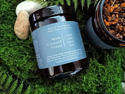 A dark amber glass jar of Root to Root Herbal Wild Placer Collection 'Moss Covered Forest' herbal tea, styled on green moss next to a stone and an open jar showing the loose leaf tea inside.