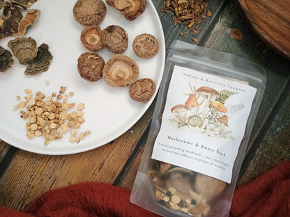 A clear stand-up pouch labeled 'Mushrooms & Roots Blend' sits on a rustic wooden surface beside a white plate holding dried mushrooms and loose herbal pieces. The pouch contains brown dried mushrooms, and the scene is styled with natural ingredients and a red cloth.