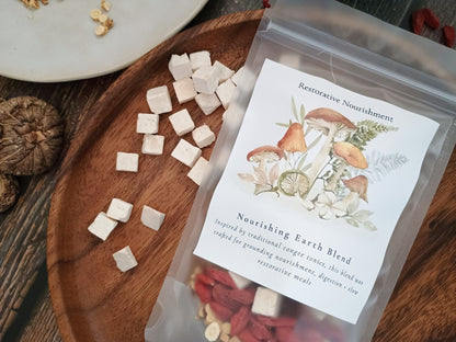 A clear stand-up pouch labeled 'Nourishing Earth Blend' restorative Nourishment, containing small white and red herbal pieces, placed on a wooden tray with loose herbal cubes nearby on a rustic wood surface.
