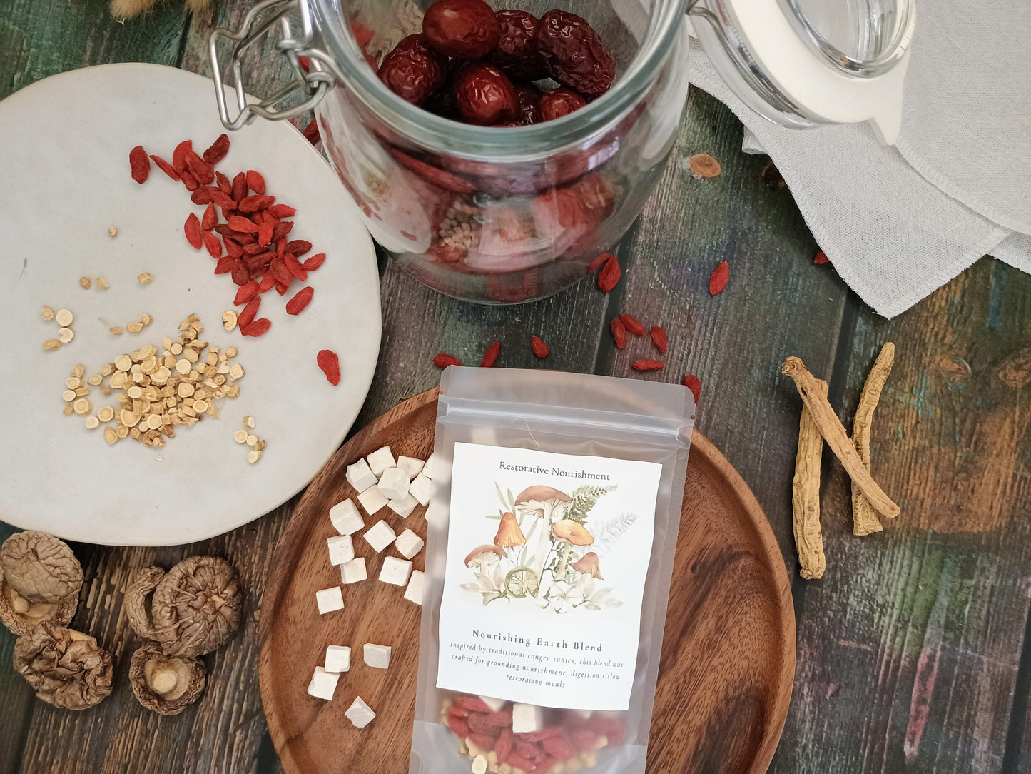 A clear pouch labeled 'Nourishing Earth Blend' containing dried herbs and red goji berries, arranged on a rustic wooden surface with loose ingredients including red goji berries, beige seeds, white poria cubes, and brown roots around it.