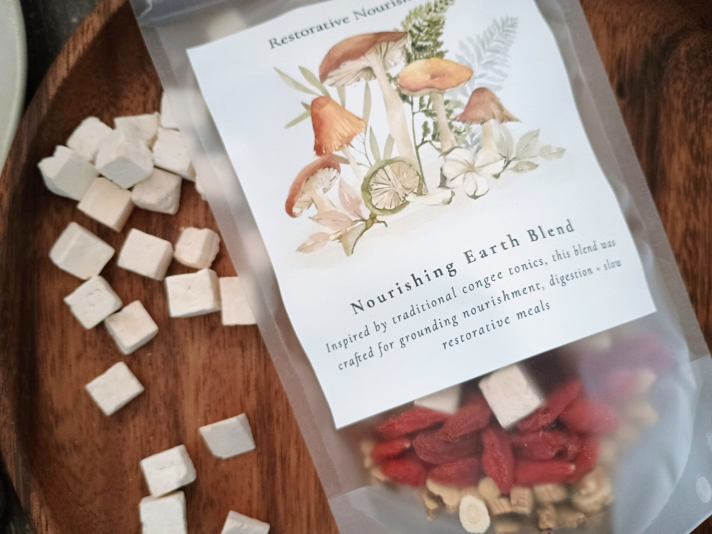 A clear stand-up pouch labeled 'Nourishing Earth Blend' containing herbal tea or tonic ingredients, placed on a dark wooden tray with scattered white cube-shaped pieces around it. The label shows illustrated mushrooms, flowers, and leaves.