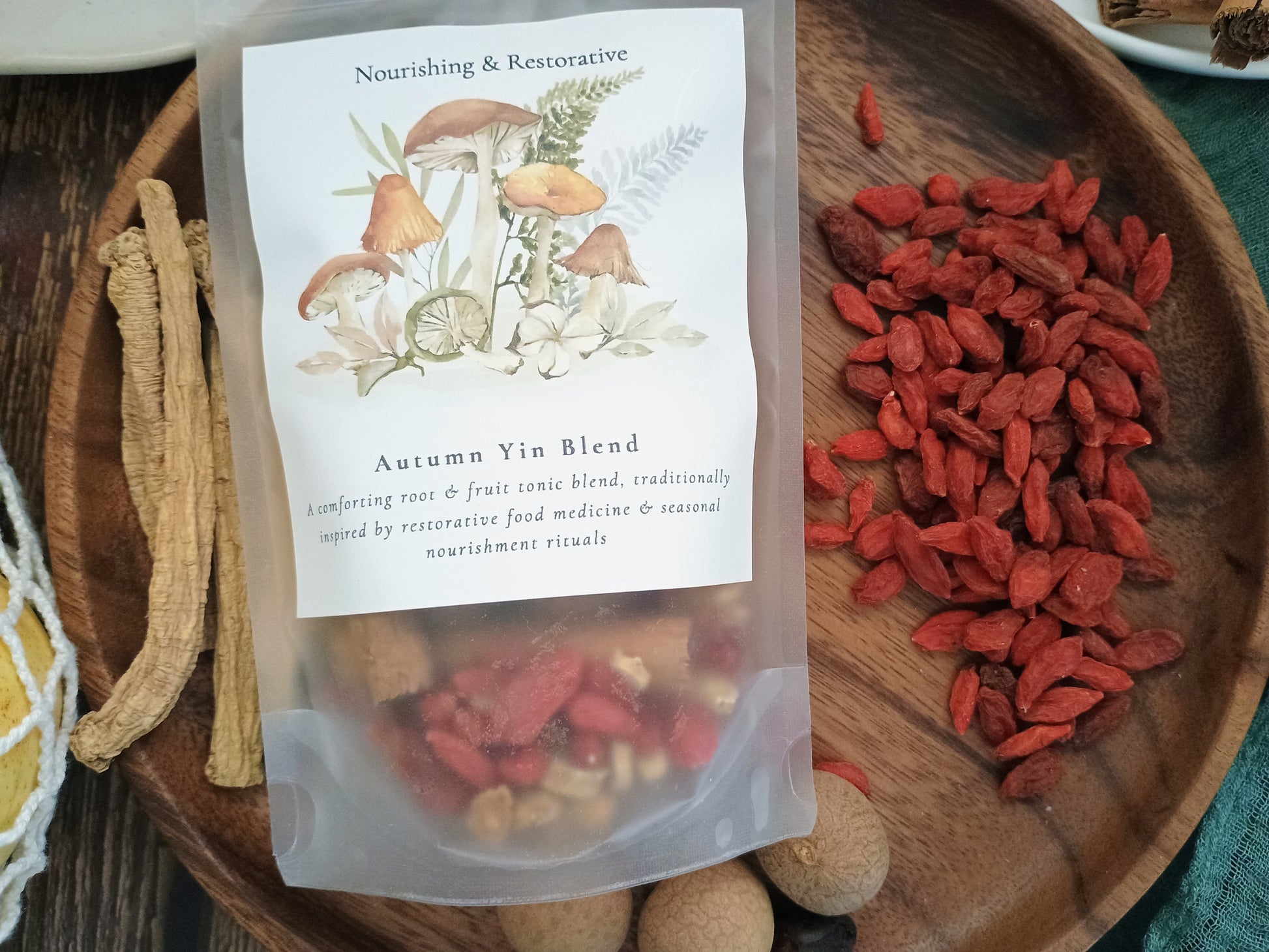 A clear stand-up pouch labeled 'Autumn Yin Blend' by Nourishing & Restorative, placed on a wooden tray beside loose red goji berries and dried herbal pieces. The pouch has a white botanical label with mushroom and leaf illustrations.