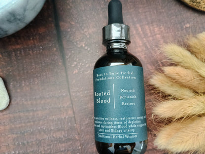 A dark amber glass dropper bottle labeled 'Rooted Blood' from the 'Root to Bone Herbal foundations Collection', placed on a dark wooden surface with dried grass and stones as props.