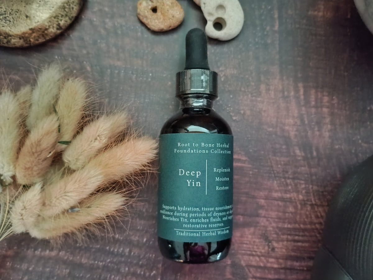 A dark amber glass dropper bottle labeled 'Deep Yin' by Root to Bone Herbal Formulas Collection, placed on a dark wooden surface next to dried bunny tail grass and small natural stones.