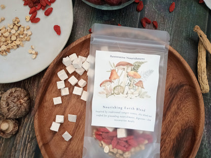 A clear stand-up pouch labeled 'Nourishing Earth Blend' restorative Nourishment, filled with dried red goji berries and light tan ingredients, displayed on a wooden tray with scattered white cubes and dried herbs around it.