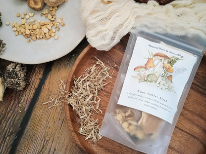 A clear stand-up pouch labeled 'Root Cellar Blend' with mushroom illustrations, placed on a wooden tray beside loose dried herbs and a white cloth on a rustic wood surface.