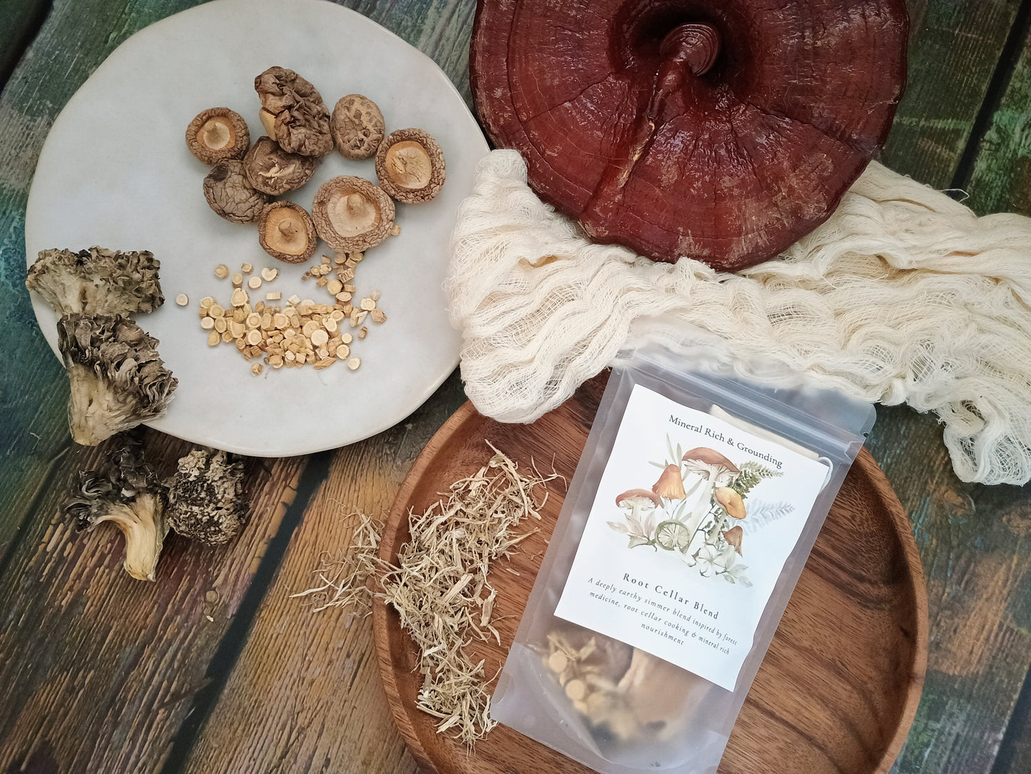 A styled flat-lay of raw mushroom products on a rustic wooden table, featuring a clear pouch of raw celery and mushroom blend, a large red reishi mushroom, sliced shiitake mushrooms, dried morels, and herbal pieces on a white plate and wooden tray.