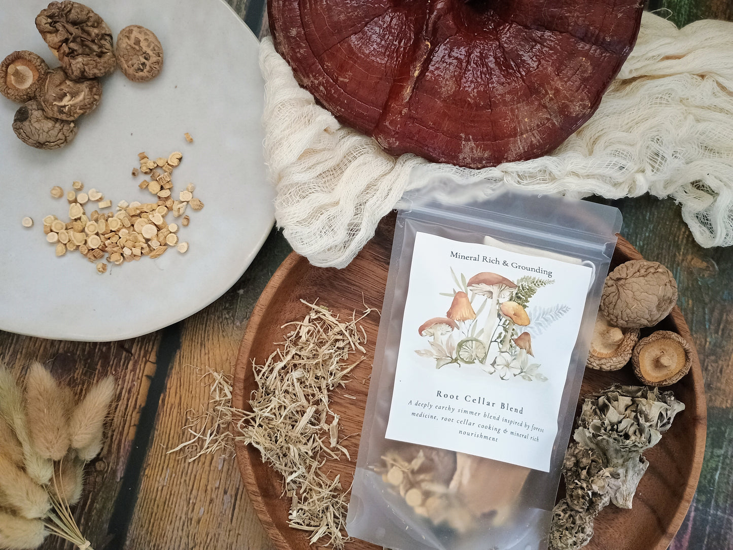 A clear stand-up pouch labeled 'Root Cellar Blend' with mushroom illustrations, styled on a wooden surface with dried herbs, mushrooms, seeds, cloth, and a large reddish-brown root vegetable.
