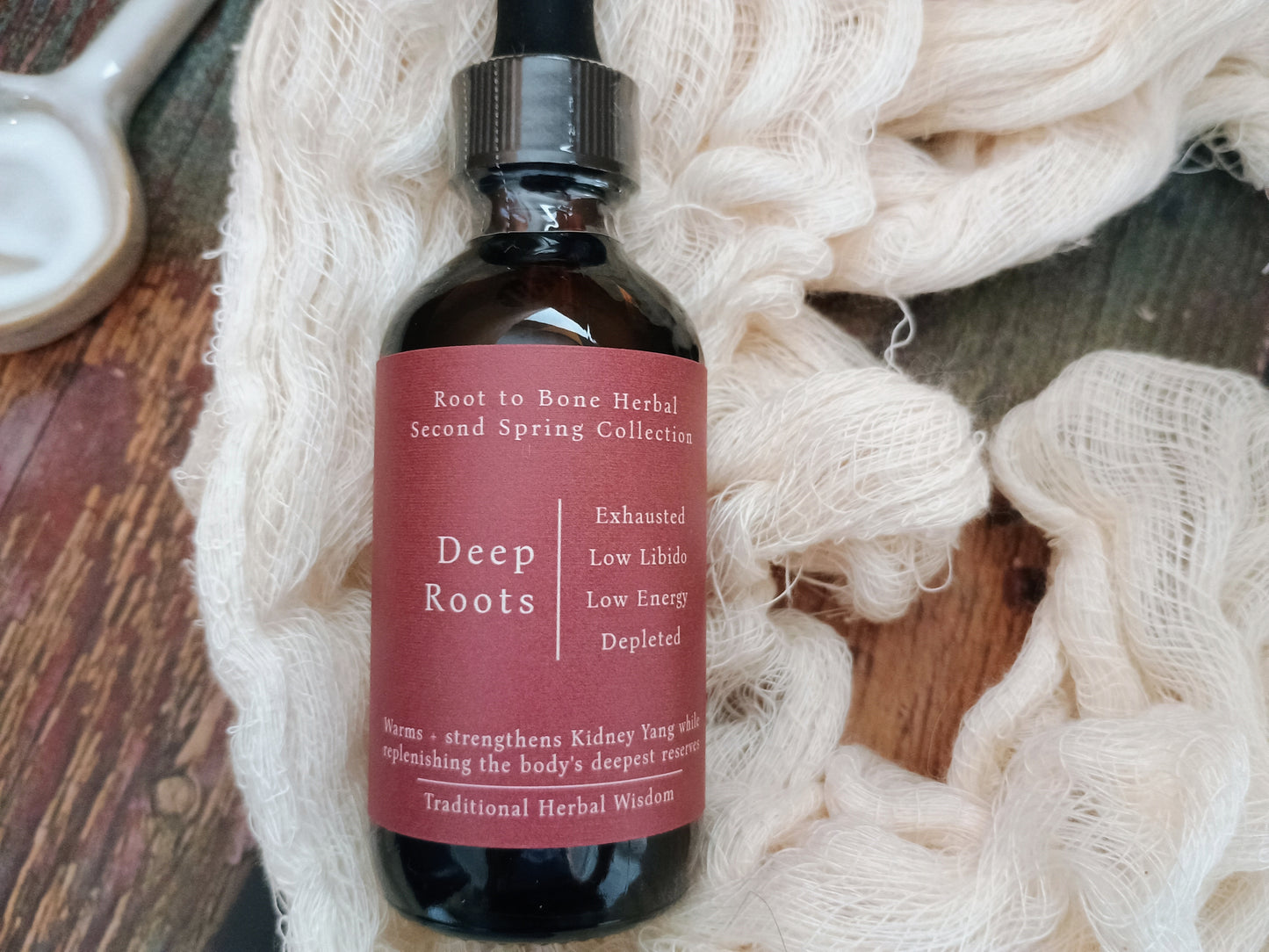An amber glass dropper bottle labeled 'Deep Roots' by Root to Bone Herbal, styled on a rustic wooden surface with white nettle leaves and a white ceramic spoon.