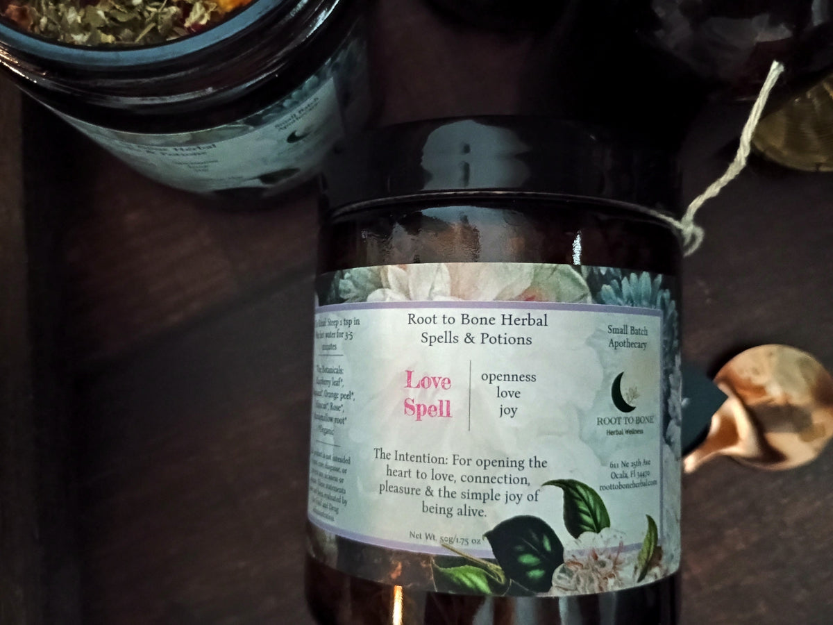 A dark glass jar candle labeled 'Love Spell' by Root to Bone Herbal Spells & Potions, placed on a dark wooden surface next to other apothecary jars.