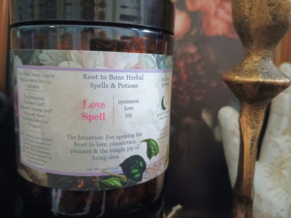 A close-up of a dark glass jar labeled 'Root to Bone Herbal Spells & Potions' with a white label featuring pink text and green leaf illustrations, set against a softly blurred background with a metallic object on the right.