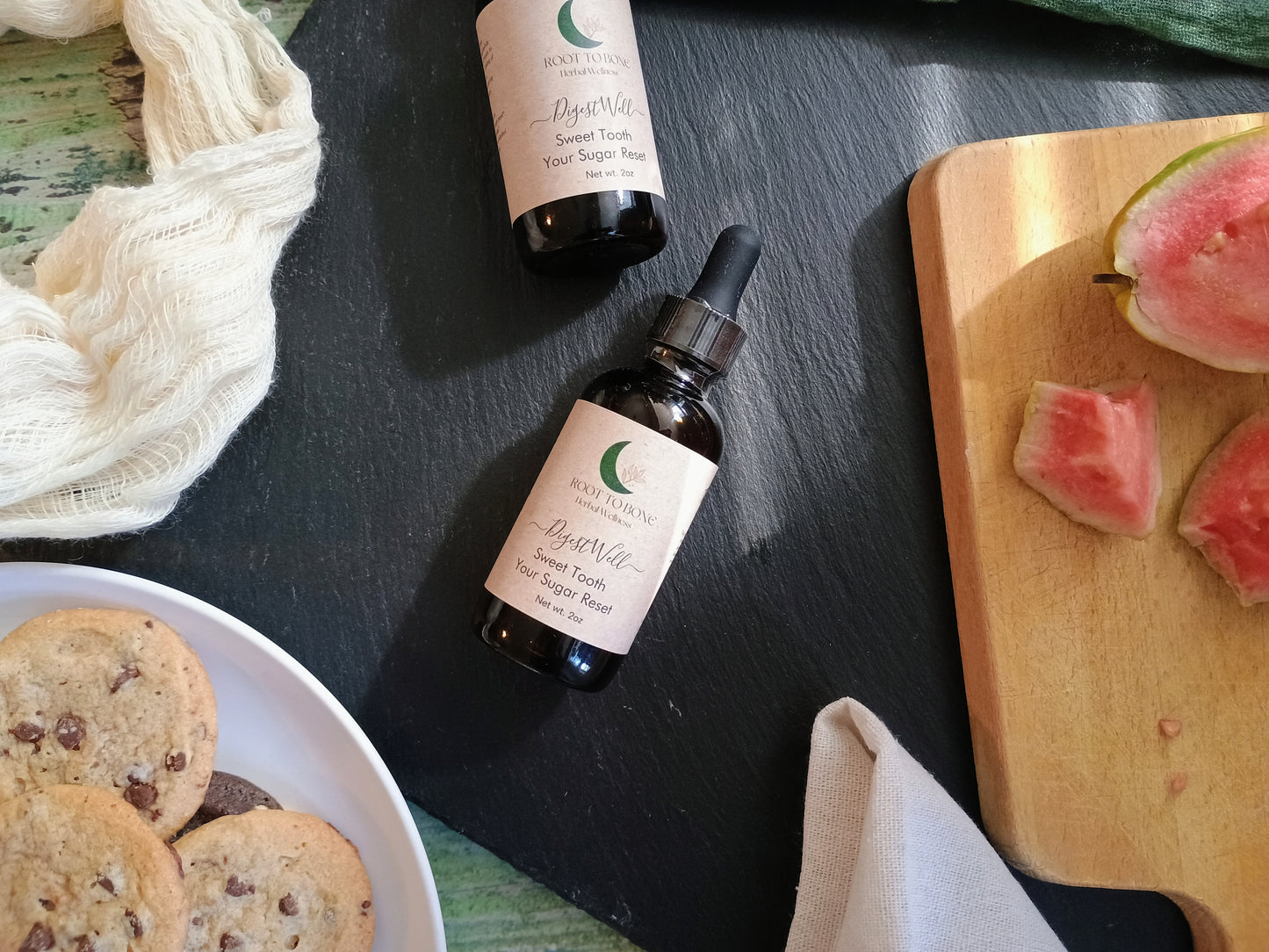 Two bottles of 'Root to Bone Herbal' on a dark surface with cookies and watermelon slices.