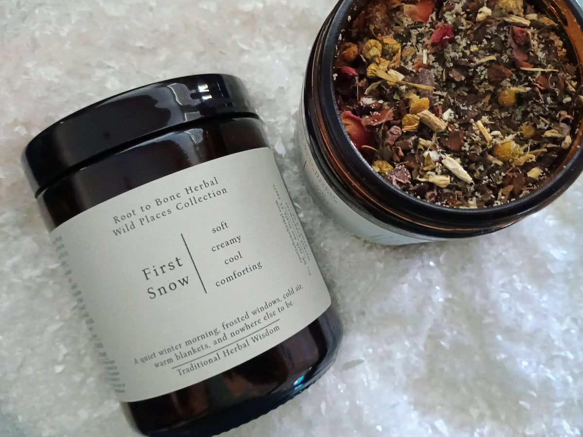 An amber glass jar of First Snow Root to Bone Herbal balm next to an open jar showing the dried herbal blend inside, set on a white textured background.