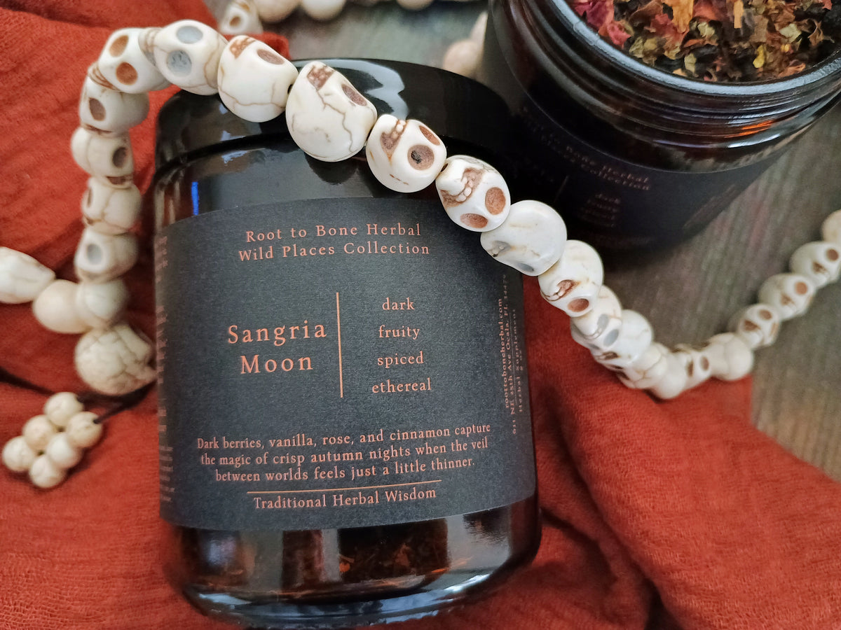 A dark amber glass jar of Root to Bone Herbal Wild Places Collection 'Sangria Moon' herbal tea, styled with a white and brown marbled bead necklace on a rust-orange fabric background.