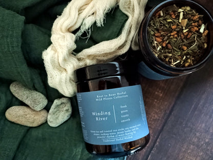 A dark amber herbal tea jar labeled 'Winding River' by Root in Root Herbal, styled on a dark wood surface with a green cloth, cream-colored netting, and a small dish of loose herbal tea.
