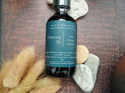A dark amber glass bottle of Root to Bone Herbal Foundations Collection Flowing Qi tincture styled on a dark wood background with decorative stones and dried grass.