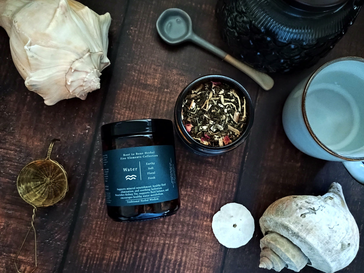 A dark glass jar labeled 'Water' with a navy label, styled on a dark wooden surface with a loose-leaf tea bowl, metal scoop, ceramic mug, and seashells.