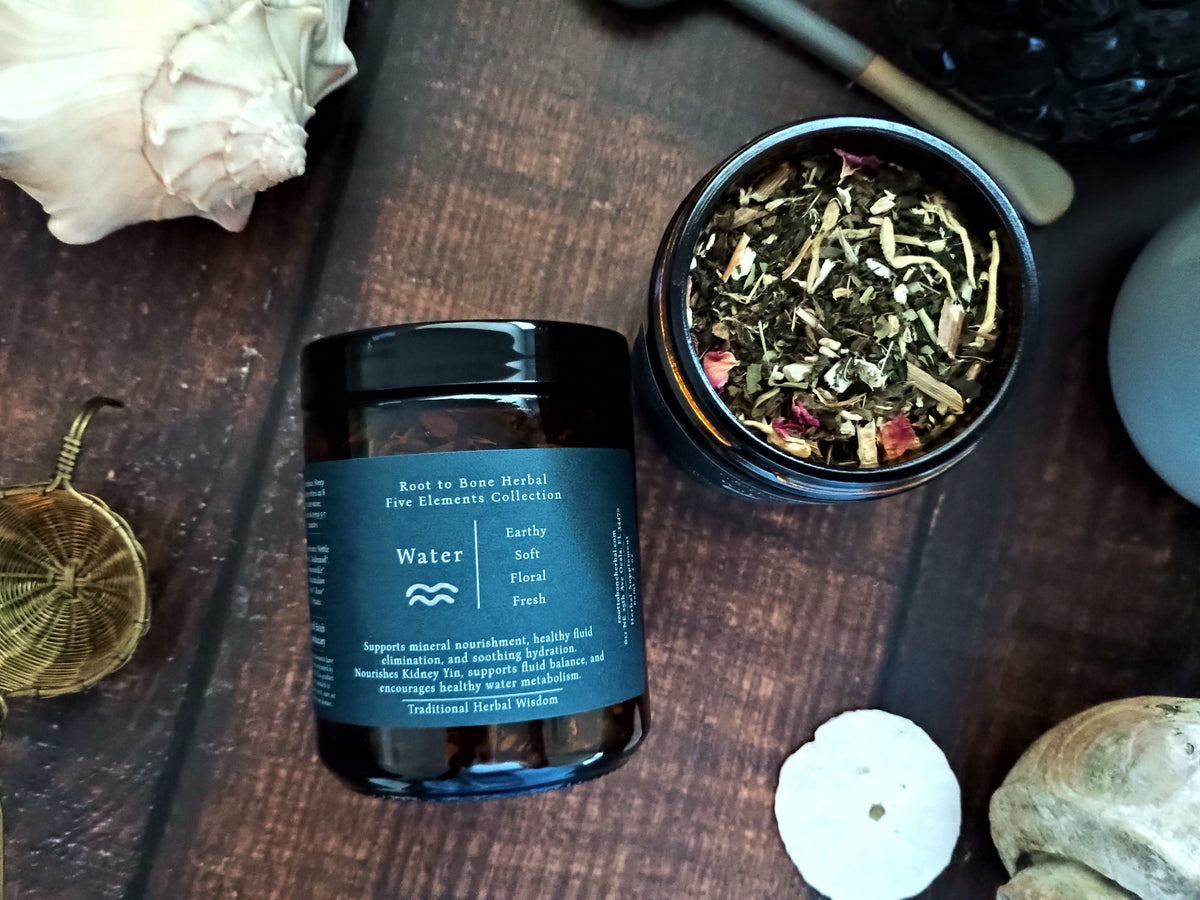 A dark amber glass jar of Root to Bone Herbal Five Elements Collection 'Water' tea, shown next to an open jar displaying the dried herbal blend on a dark wooden background with natural props