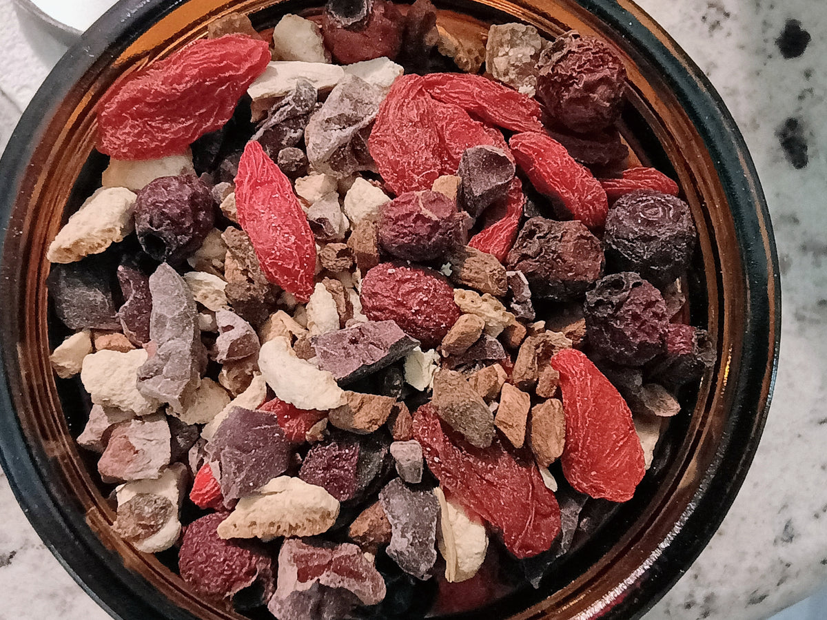 A close-up top-down view of a dark brown ceramic bowl filled with a colorful herbal tea blend containing red goji berries, dried purple fruits, brown bark pieces, and light beige roots on a speckled white countertop.