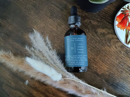 A dark amber glass dropper bottle labeled 'Yi' by Root to Bone Herbs, styled on a rustic wooden surface with dried pampas grass and a decorative plate in the background.